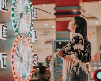 A woman is standing in an arcade looking at a large colorful game machine with bright lights. The game machine features large spinning wheels labeled with 'tickets' and has a vibrant design. The woman is casually dressed in a jacket with fringe details and is holding something in her hand while wearing a smartwatch.